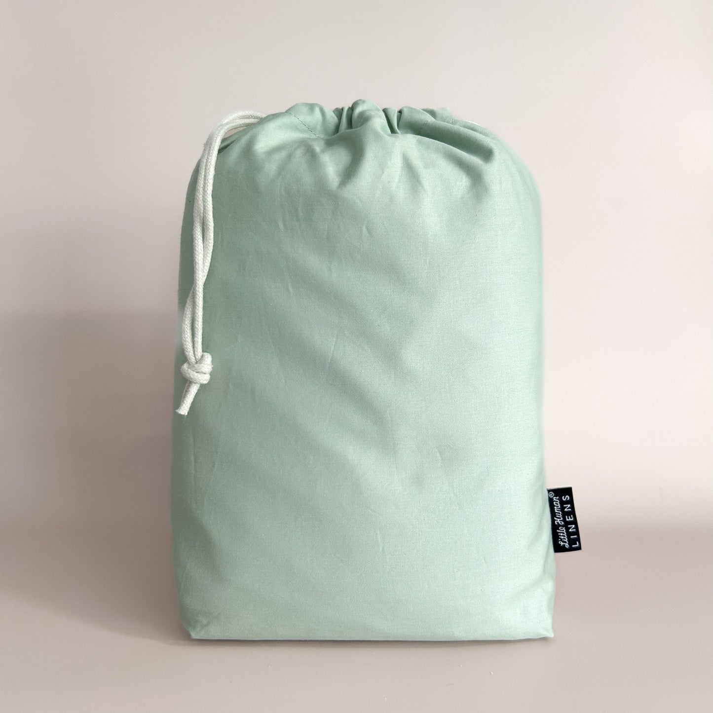A sage green European cot / crib waterproof fitted sheet packaged in a drawstring bag with a label showing the Little Human Linens brand logo.