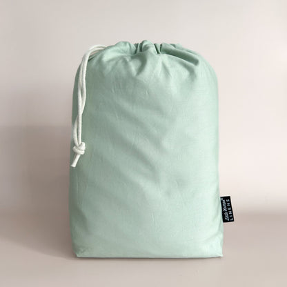 A sage green European cot / crib waterproof fitted sheet packaged in a drawstring bag with a label showing the Little Human Linens brand logo.