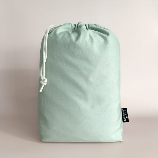 A sage green European cot / crib waterproof fitted sheet packaged in a drawstring bag with a label showing the Little Human Linens brand logo.