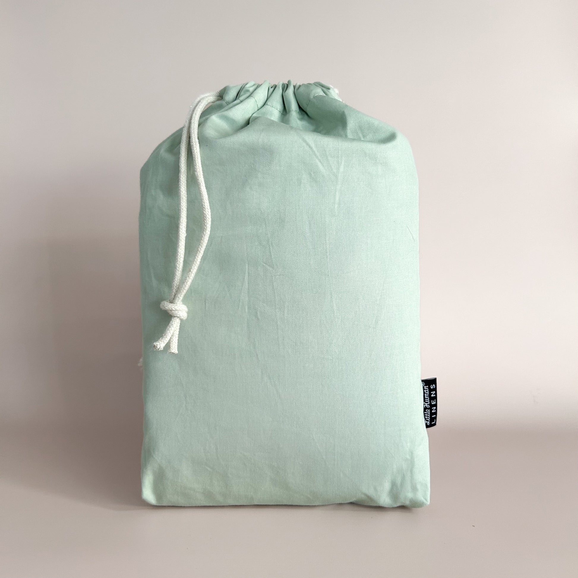 A sage green mini or compact cot / crib waterproof fitted sheet packaged in a drawstring bag with a label showing the Little Human Linens brand logo.
