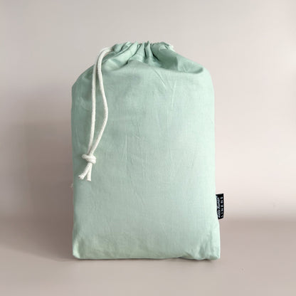 A sage green mini or compact cot / crib waterproof fitted sheet packaged in a drawstring bag with a label showing the Little Human Linens brand logo.