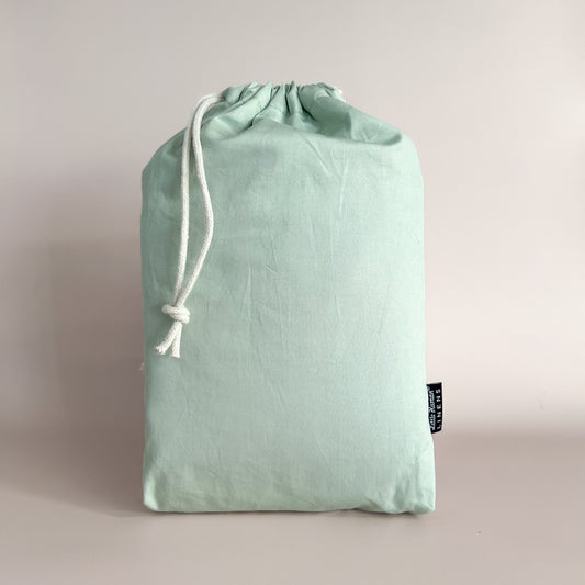 A sage green mini or compact cot / crib waterproof fitted sheet packaged in a drawstring bag with a label showing the Little Human Linens brand logo.