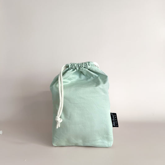 A sage green small bassinet waterproof fitted sheet packaged in a drawstring bag with a label showing the Little Human Linens brand logo.