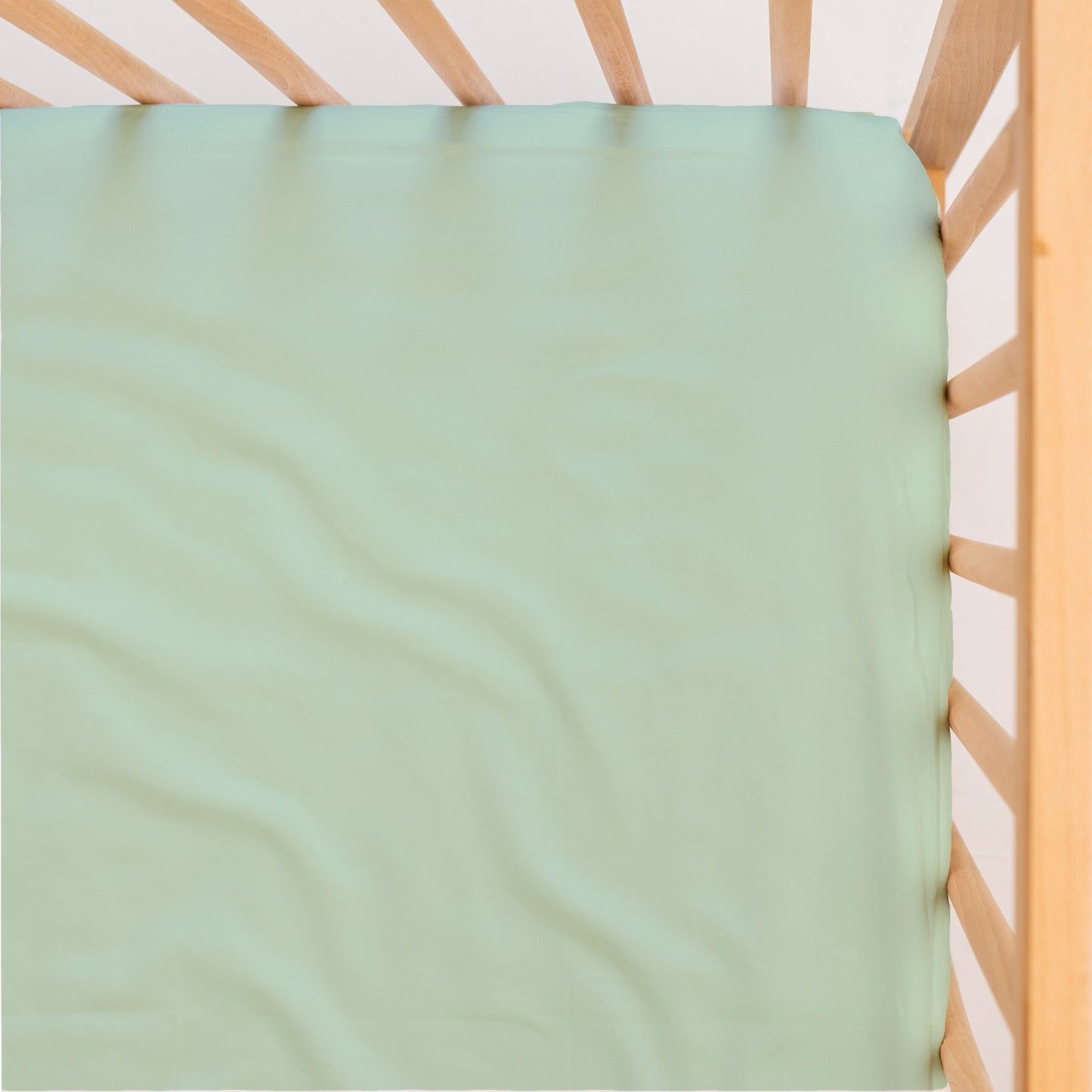 Sage green cot/crib waterproof sheet on a wooden crib with a white background, viewed from above