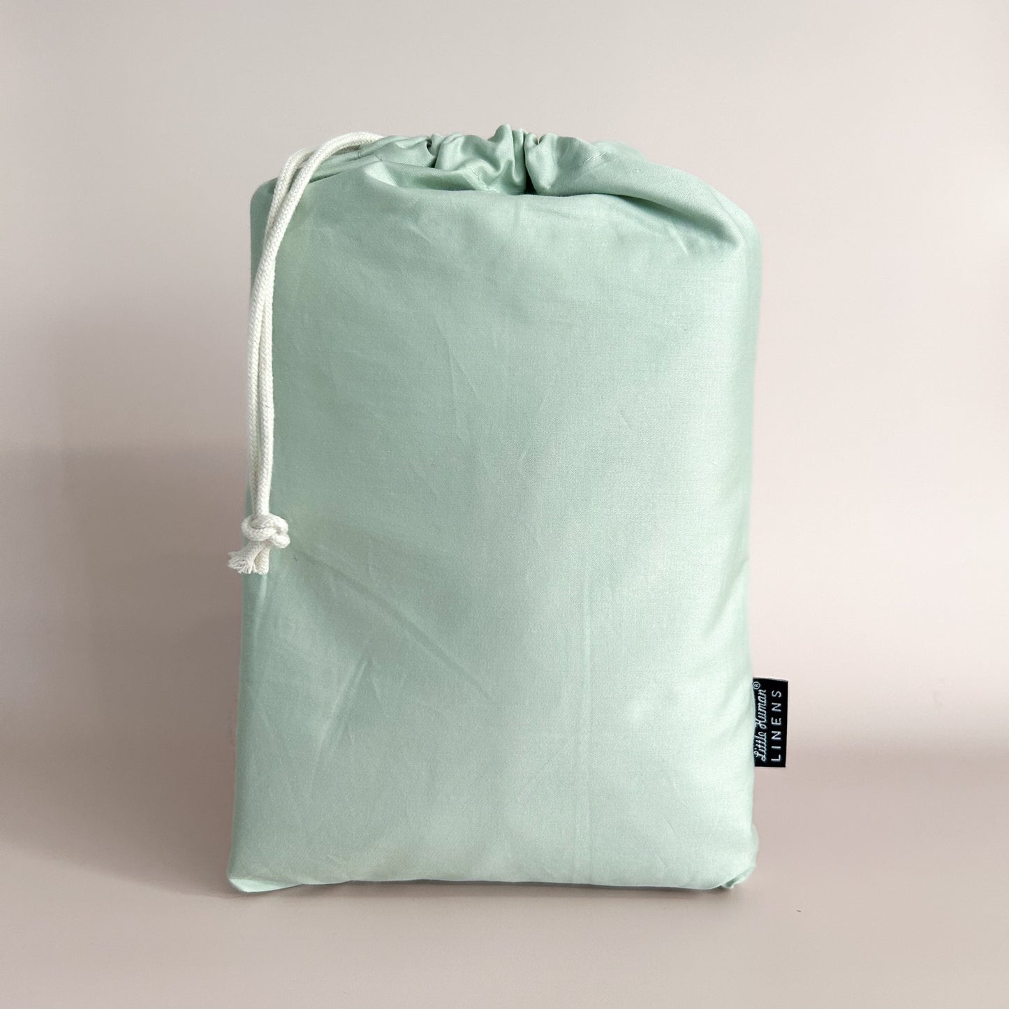 A sage green cot / crib waterproof fitted sheet packaged in a drawstring bag with a label showing the Little Human Linens brand logo.