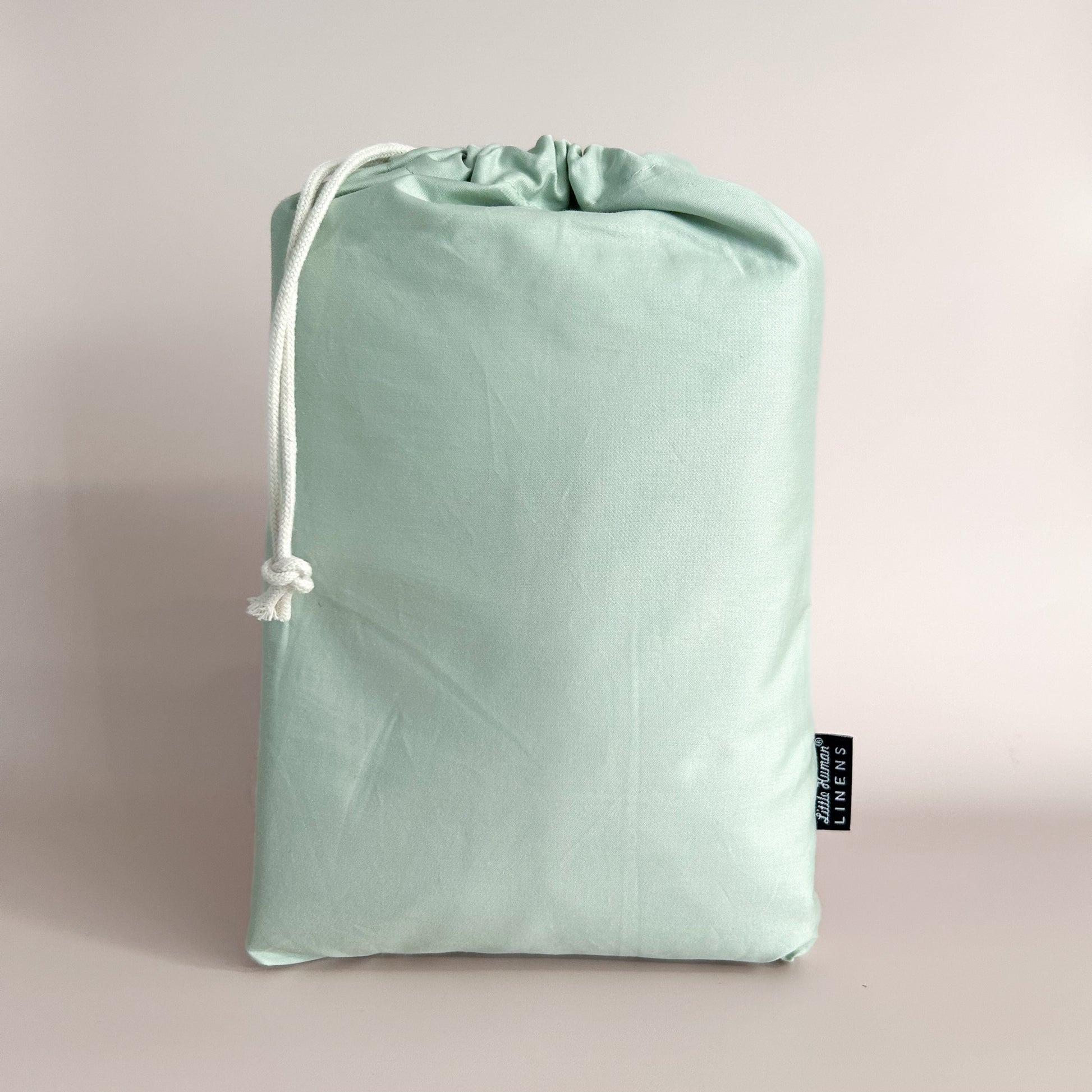 A sage green cot / crib waterproof fitted sheet packaged in a drawstring bag with a label showing the Little Human Linens brand logo.