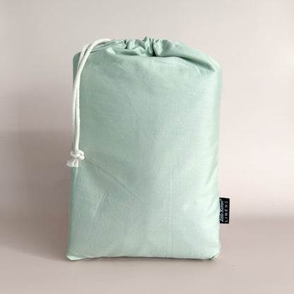 A sage green cot / crib waterproof fitted sheet packaged in a drawstring bag with a label showing the Little Human Linens brand logo.